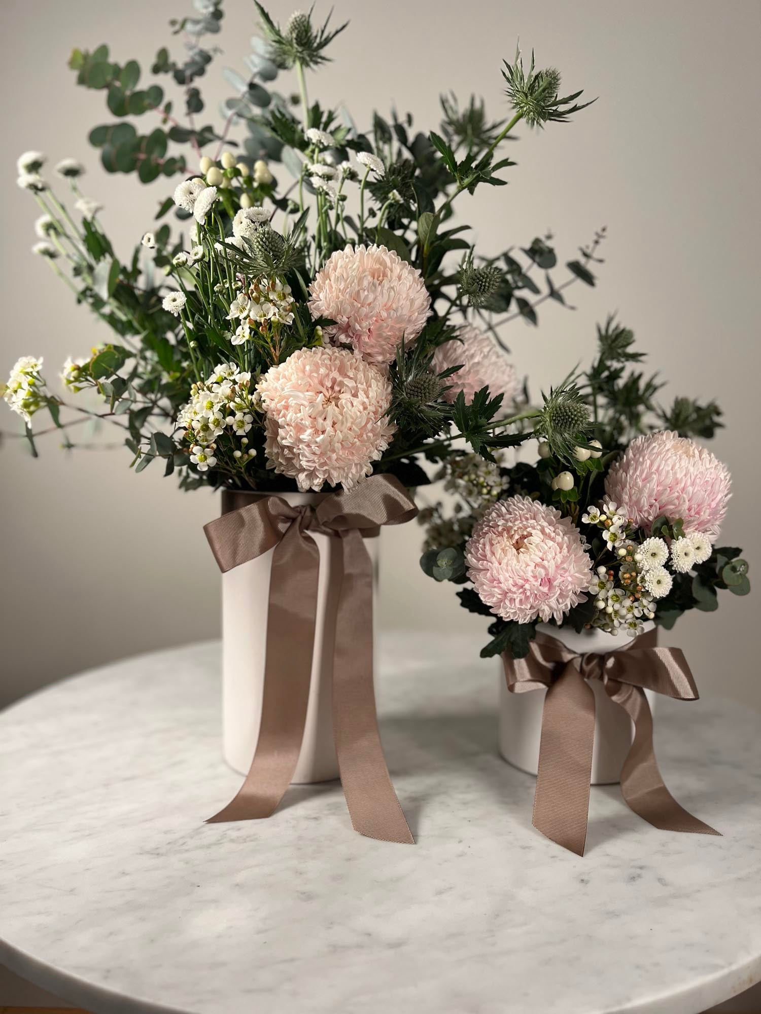 Two flower arrangements in white pots with brown ribbons on a marble surface.