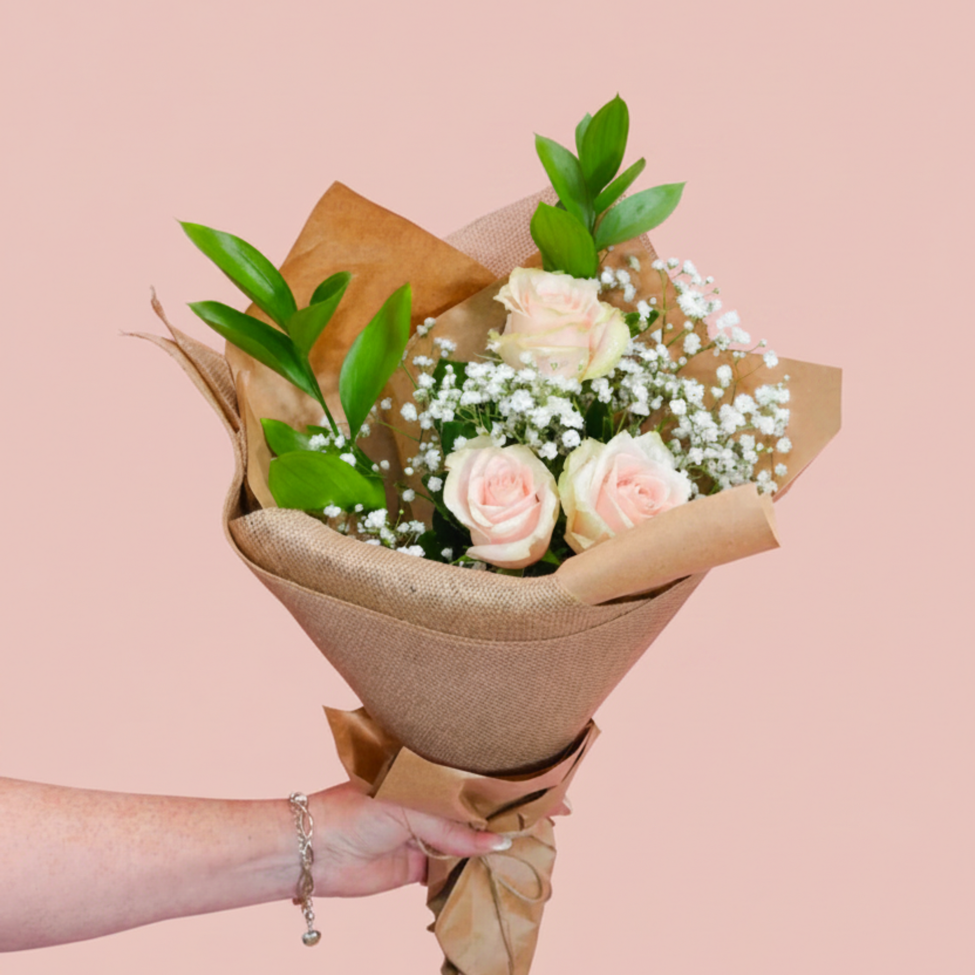 A hand holding a bouquet of three pale pink roses, white baby's breath, and green leaves, wrapped in brown kraft paper against a solid dusty pink background.