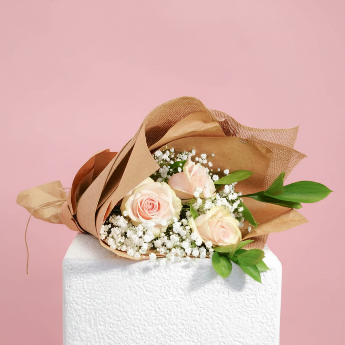 A bouquet of three pale pink roses, white baby's breath, and green leaves, wrapped in brown kraft paper, displayed on a white pedestal against a solid pink background.