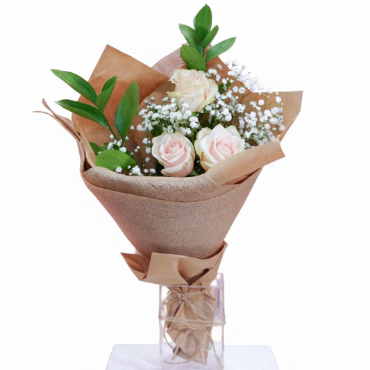 A floral bouquet with three pale pink roses, white baby's breath, and green leaves, wrapped in brown kraft paper and displayed in a clear square glass vase on a white pedestal against a white background.