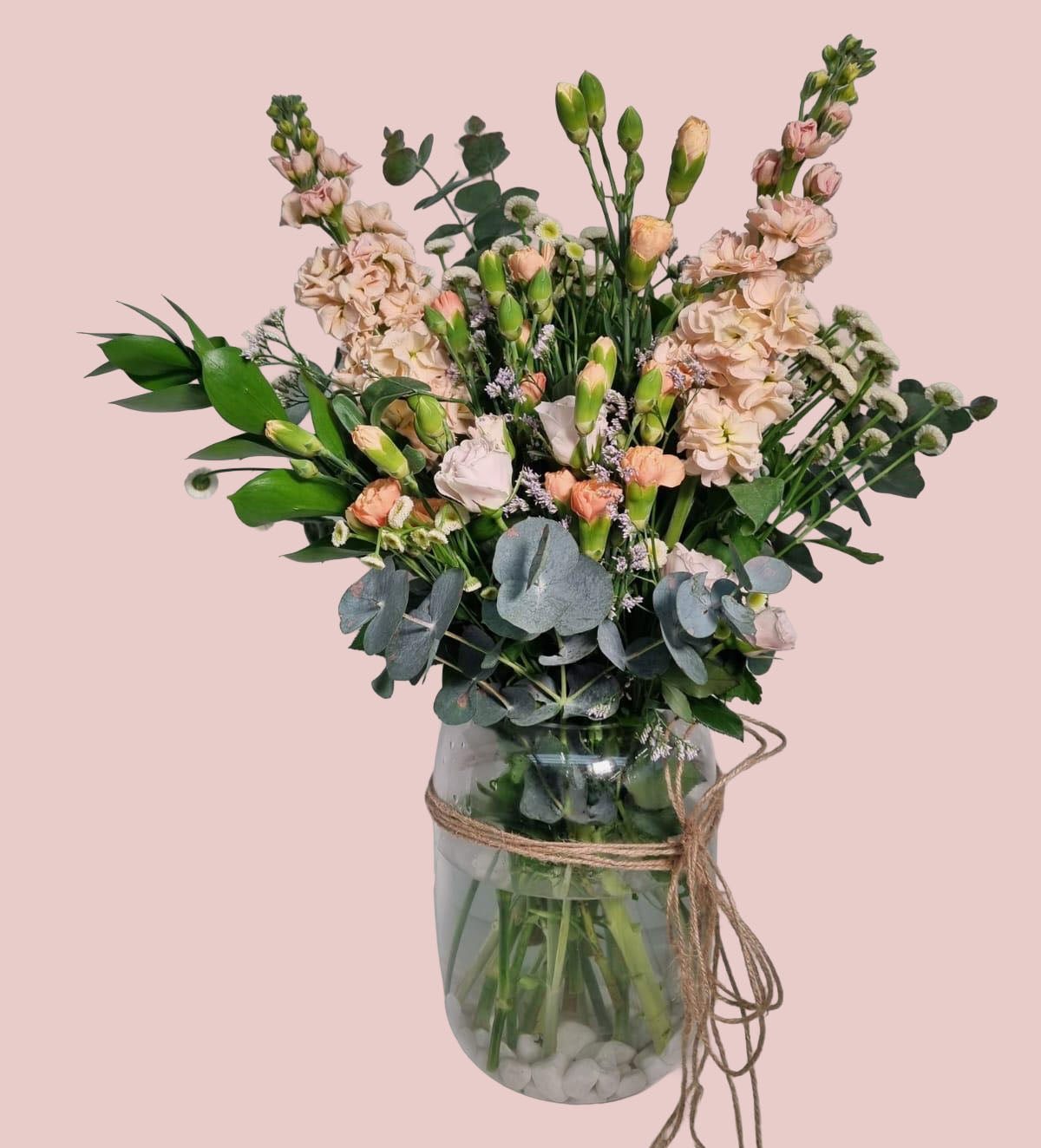 A floral arrangement featuring blush pink flowers, green foliage, and eucalyptus leaves in a clear glass jar tied with twine, set against a solid pale pink background.