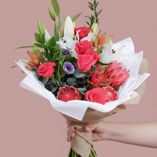 A floral bouquet wrapped in white tissue paper and a burlap band, arranged in a clear glass jar. The bouquet features bright pink roses, white lilies, green leaves, and protea flowers, set against a solid pink background.
