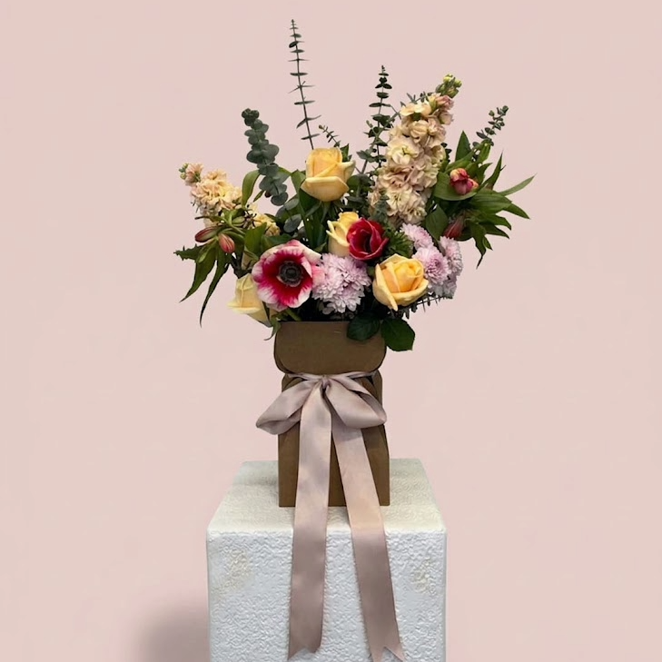A floral arrangement in a brown box with a beige ribbon, displayed on a white pedestal against a soft pink background.