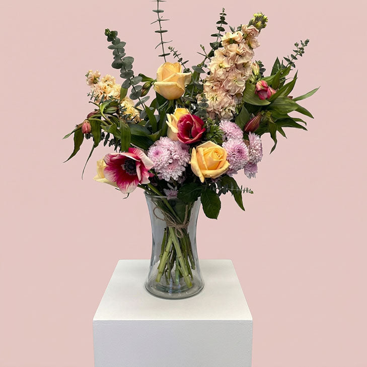 A vibrant floral arrangement featuring yellow roses, pink and red flowers, and green foliage in a clear glass vase, displayed on a white pedestal against a solid light pink background.