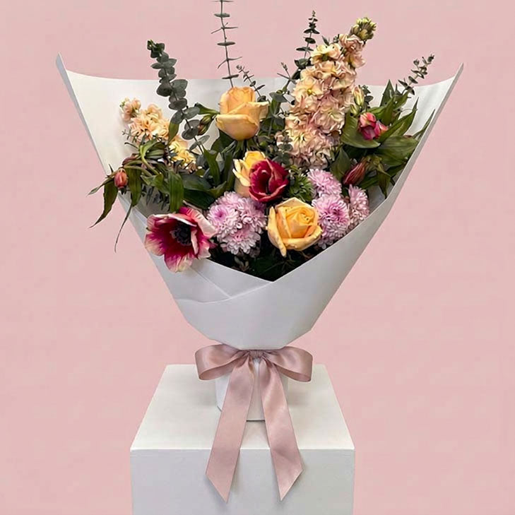 A floral bouquet wrapped in white paper and tied with a pale pink ribbon, displayed on a white pedestal against a soft pink background. The bouquet includes yellow roses, pink chrysanthemums, burgundy flowers, and green foliage.