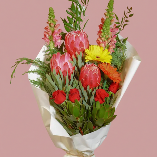 A vibrant mixed flower bouquet wrapped in white paper and presented in a light wood crate on a white pedestal, set against a solid pink background.