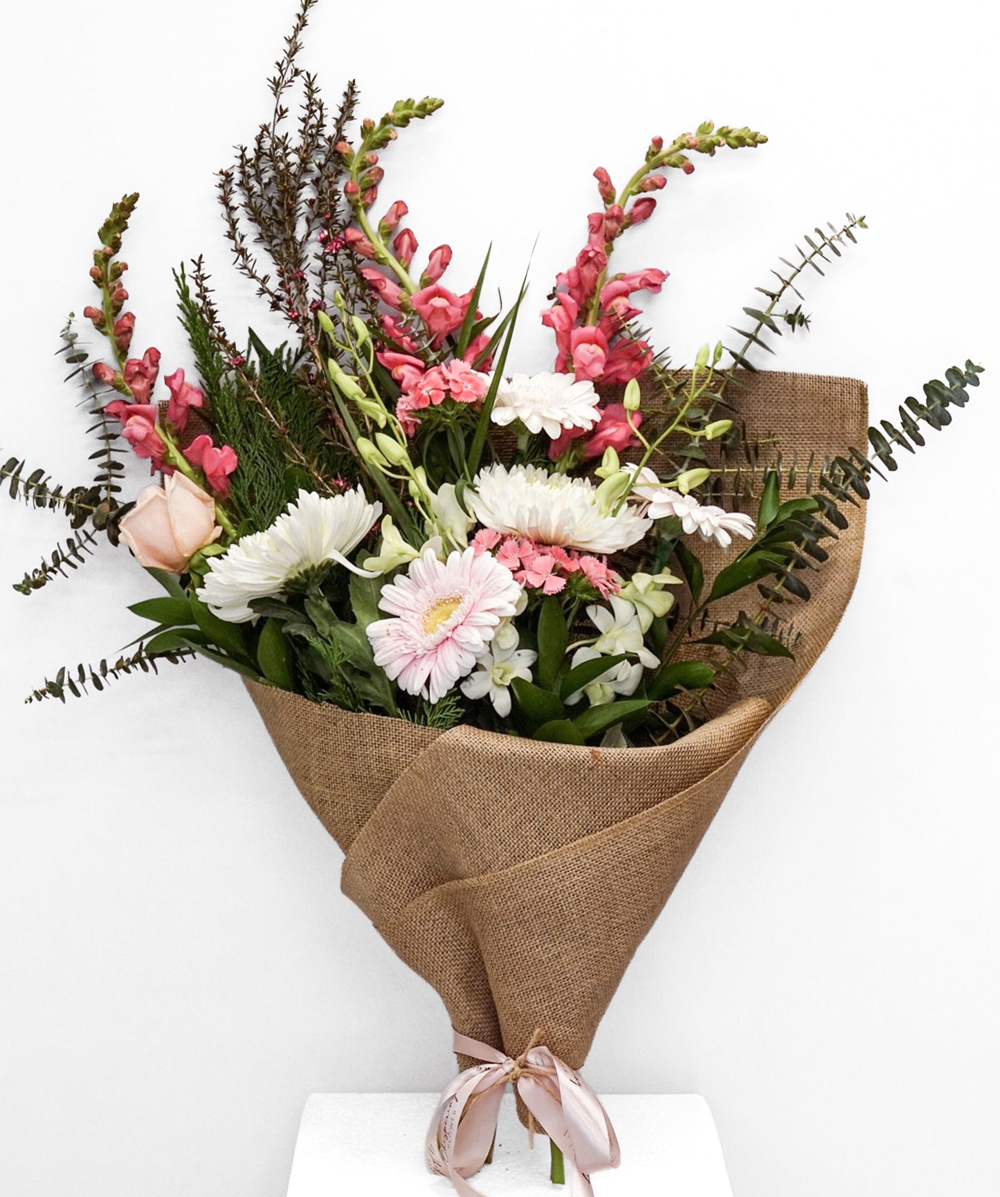 A mixed floral bouquet wrapped in brown burlap paper and tied with a pale pink ribbon, displayed on a white pedestal against a white background.