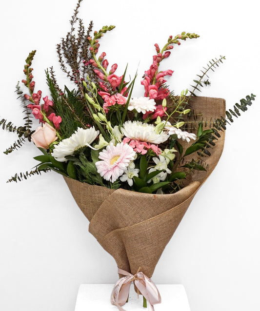 A mixed floral bouquet wrapped in brown burlap paper and tied with a pale pink ribbon, displayed on a white pedestal against a white background.