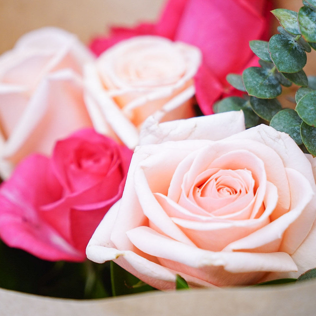 A close-up of a bouquet of roses wrapped in brown kraft paper, featuring pale peach and bright pink roses with green eucalyptus leaves.